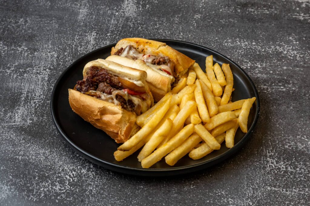 A black plate with a cheesesteak sandwich filled with beef, cheese, and onions, served alongside a portion of French fries.