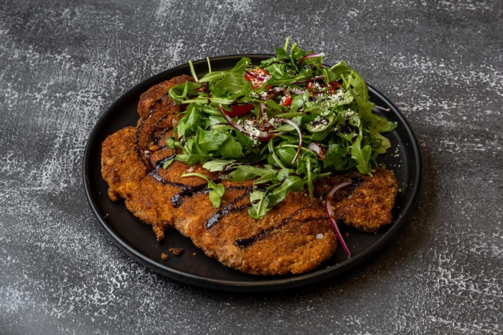 A black plate holds breaded and fried cutlets topped with fresh arugula salad, red onion, and a drizzle of dark sauce, set on a gray textured surface.
